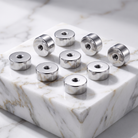 Eight silver metallic cylindrical hardware pieces with threaded holes, arranged on a white marble display block against a marble surface.