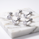 Five silver-toned labret ear piercings with clear faceted gemstones in different shapes, arranged on a white marble block against a white background.