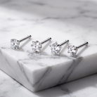 Four silver-toned stud earrings with clear gemstones in round, oval, and star shapes, arranged on a white marble block over a marble surface.