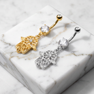 Two belly button rings with Hamsa hand charms are displayed on a white marble block. One ring is gold-toned with a clear rhinestone, and the other is silver-toned with a clear rhinestone.