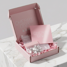 An open pink gift box labeled CHERRY DIVA sits on a white marble surface. Inside is pink crinkle paper filler, a matching pink card, and a small clear pouch containing a silver-toned decorative item.