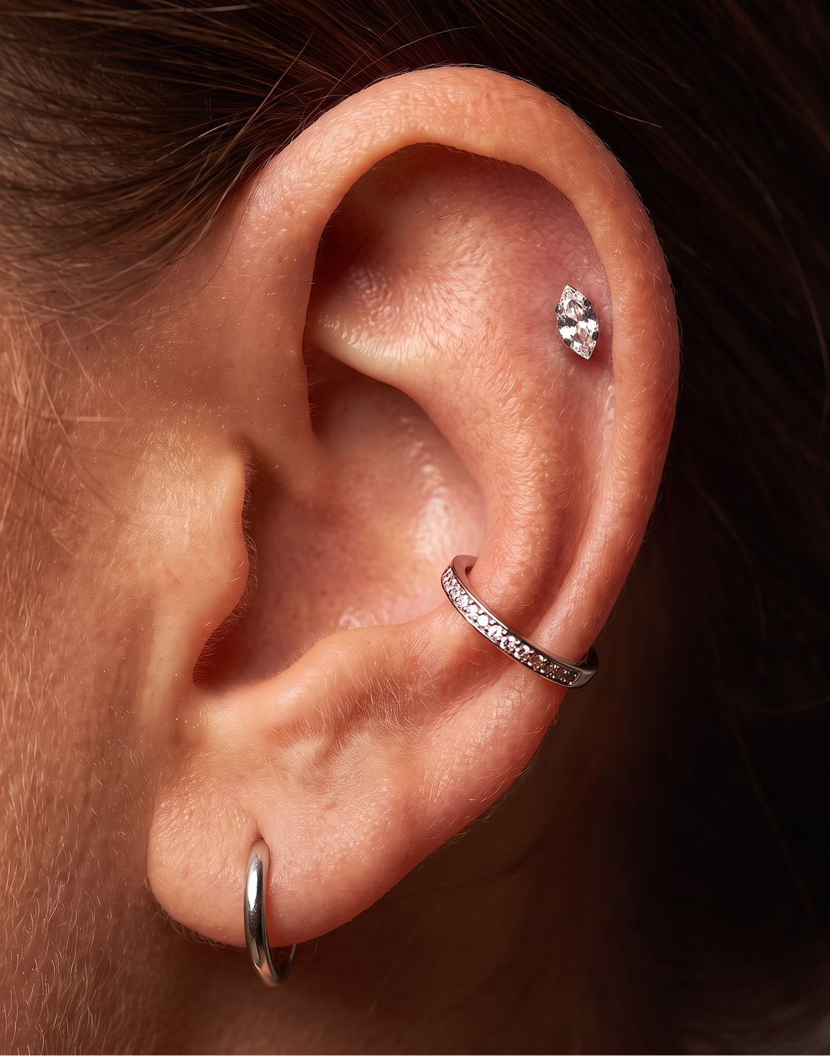 Close-up of a person's ear showcasing three silver-toned ear piercings: a small hoop in the lobe, a pavé-set huggie ring on the helix, and a small cluster stud on the lobe.