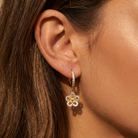 A close-up of a woman's ear wearing a gold huggie hoop earring with a dangling flower charm. The charm features a yellow center surrounded by clear stones, set against warm, golden lighting.