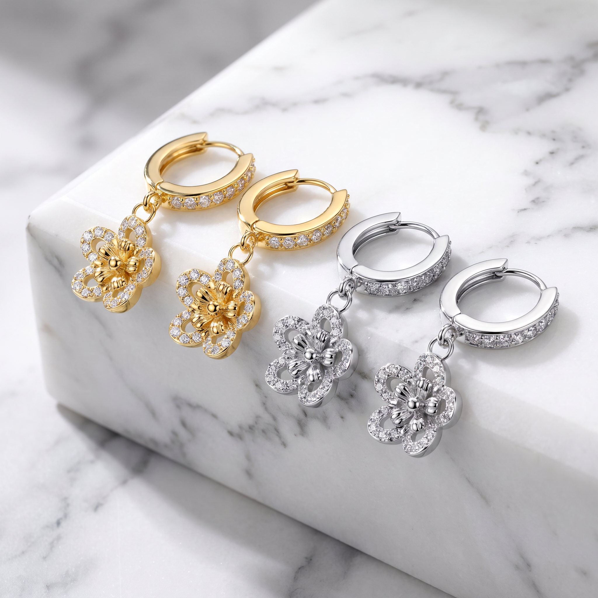 Two pairs of huggie hoop earrings with dangling flower charms, one in yellow gold and one in silver, both set with clear stones, displayed on a white marble block.