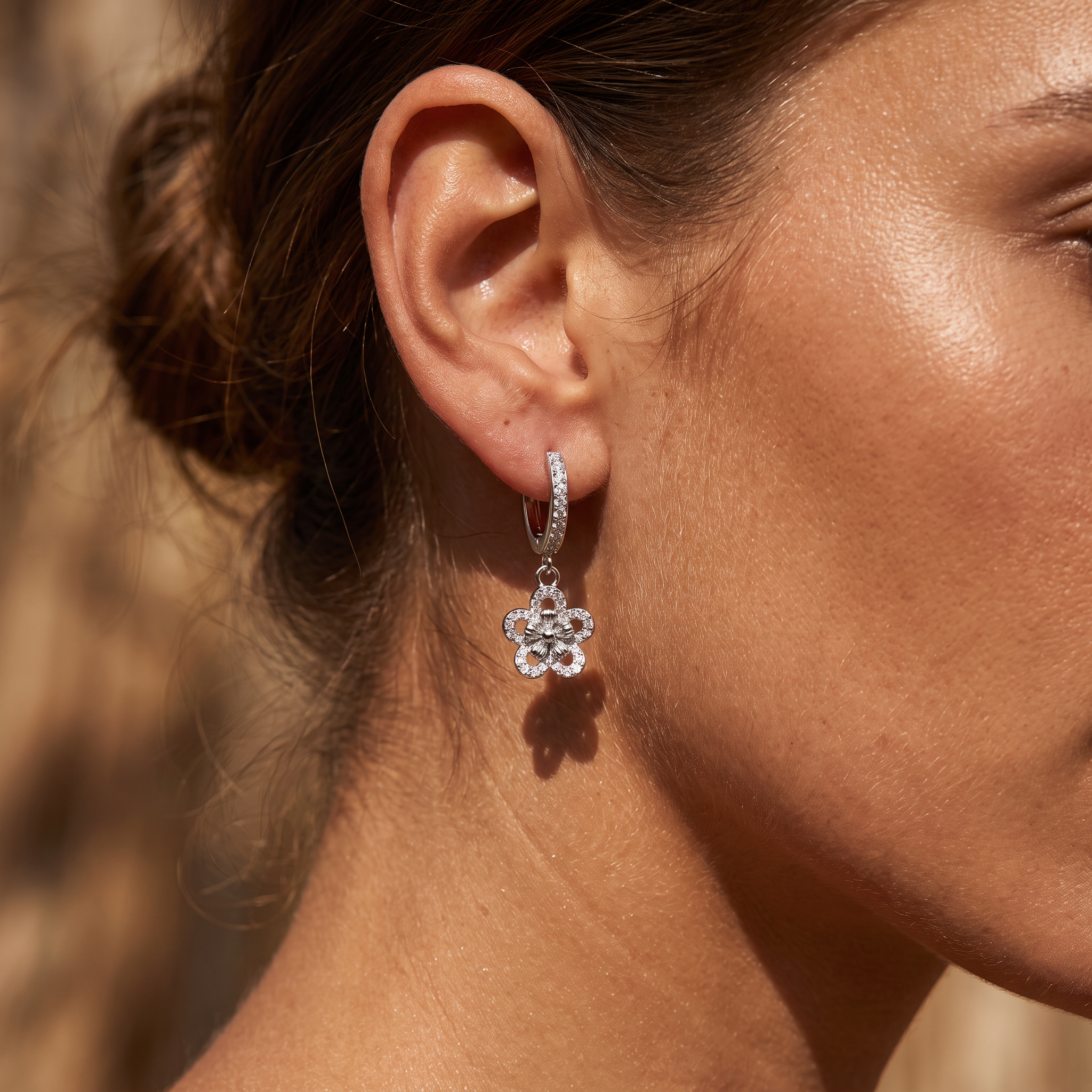A close-up of a woman's ear wearing a silver huggie hoop earring with a dangling flower-shaped crystal charm.