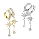 Two pairs of huggie hoop earrings with dangling star and key motifs, shown in yellow gold and silver metal finishes. The hoops and stars are set with clear pavé stones, and each dangling piece culminates in a round clear stone.