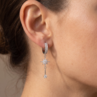 A close-up of a model's ear wearing a silver huggie hoop earring with a dangling starburst charm and key detail, set with clear gemstones.
