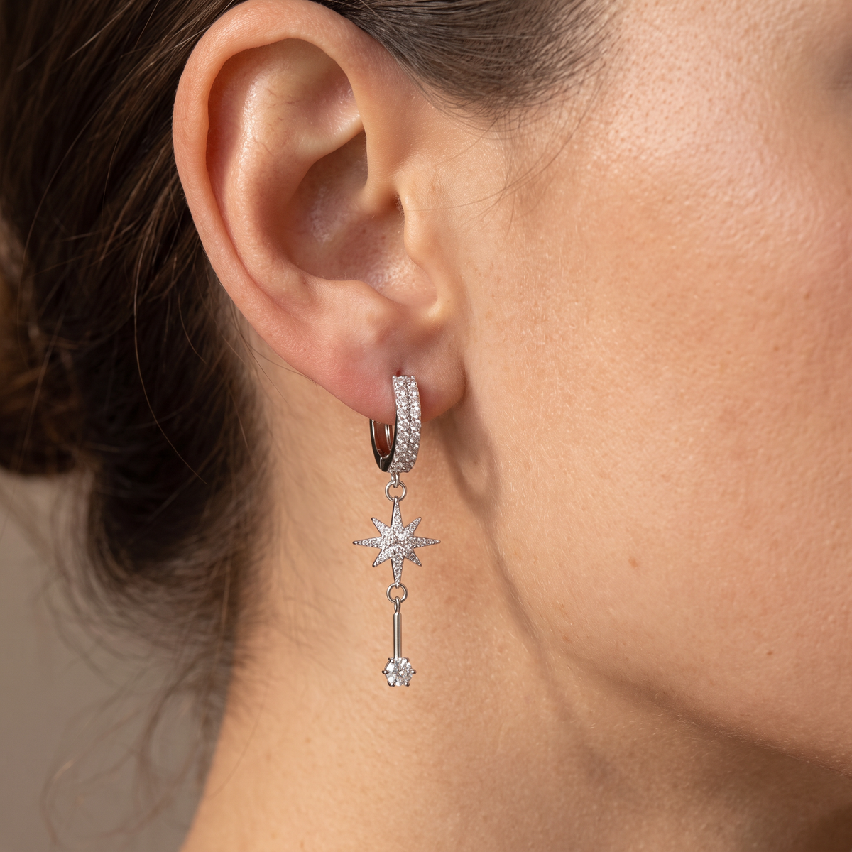 A close-up of a model's ear wearing a silver huggie hoop earring with a dangling starburst charm and key detail, set with clear gemstones.