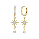 A pair of gold dangle earrings on a plain white background. One earring is a huggie hoop with a vertical bar set with clear stones, and the other is a drop earring with a starburst motif, a small link, and a round clear gemstone.
