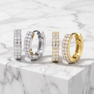 Four huggie hoop earrings displayed on a white marble slab. Two earrings are silver-toned with pavé-set clear stones, and two are gold-toned with pavé-set clear stones.