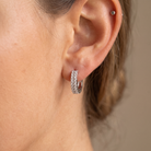 A close-up of a person's ear wearing a small silver huggie hoop earring paved with clear stones, with a tiny silver stud piercing on the upper earlobe.