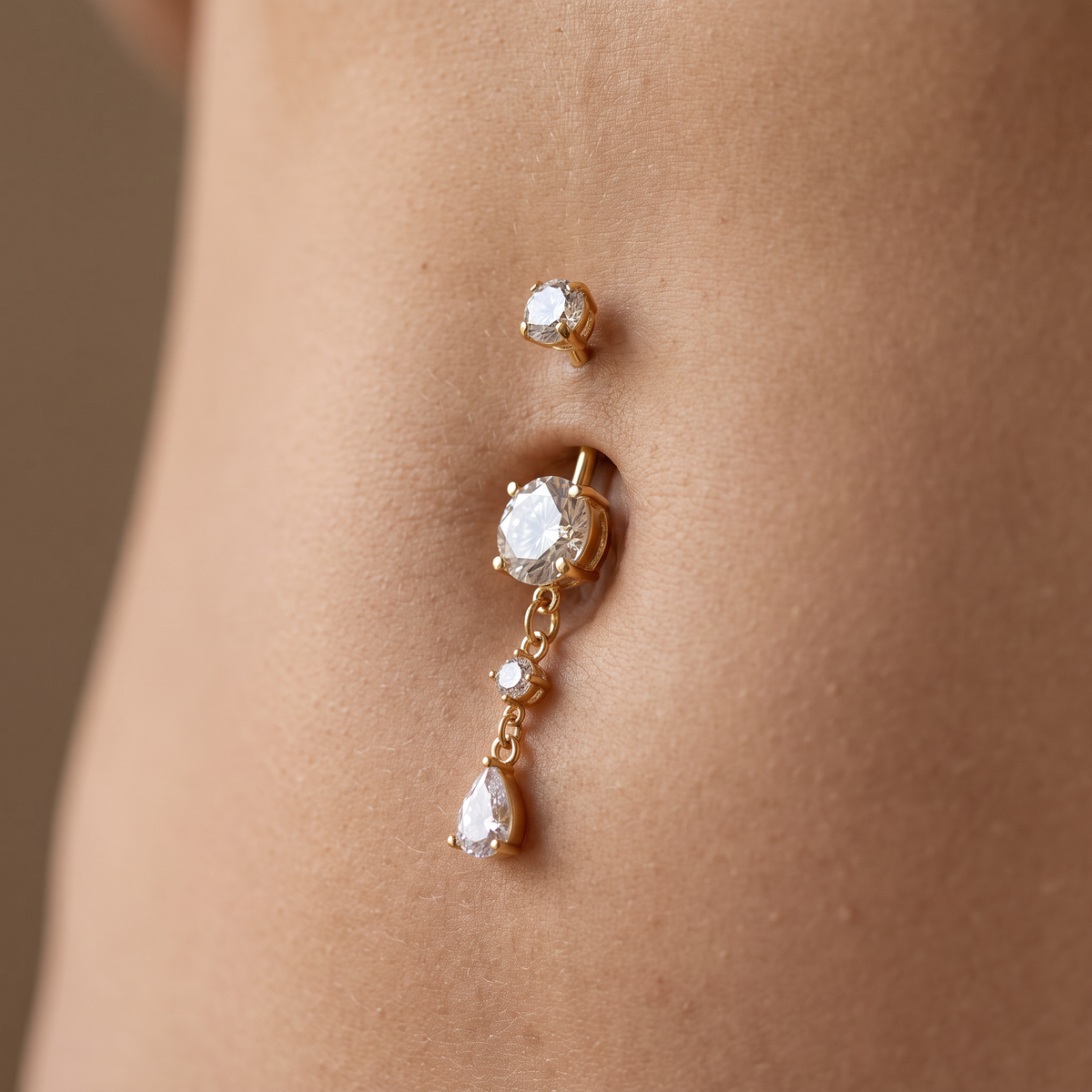 A close-up of a gold belly button ring with a teardrop dangling charm and clear gemstones, shown worn on a person's abdomen.