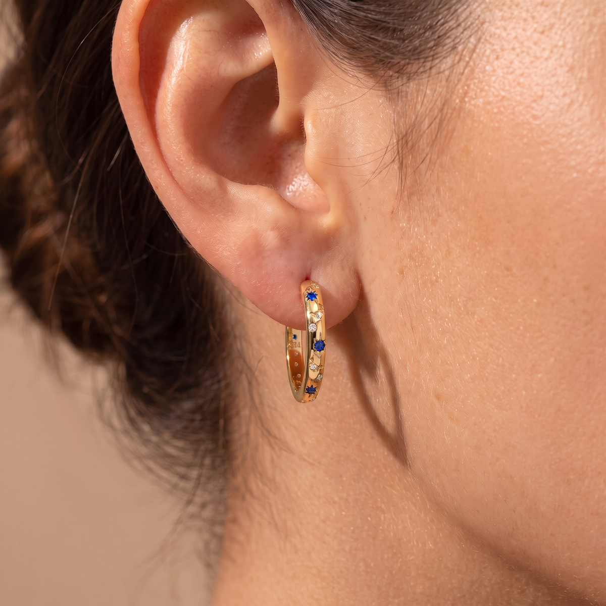 A close-up of a person's ear wearing a small gold hoop earring adorned with tiny clear stones and a single deep blue stone.