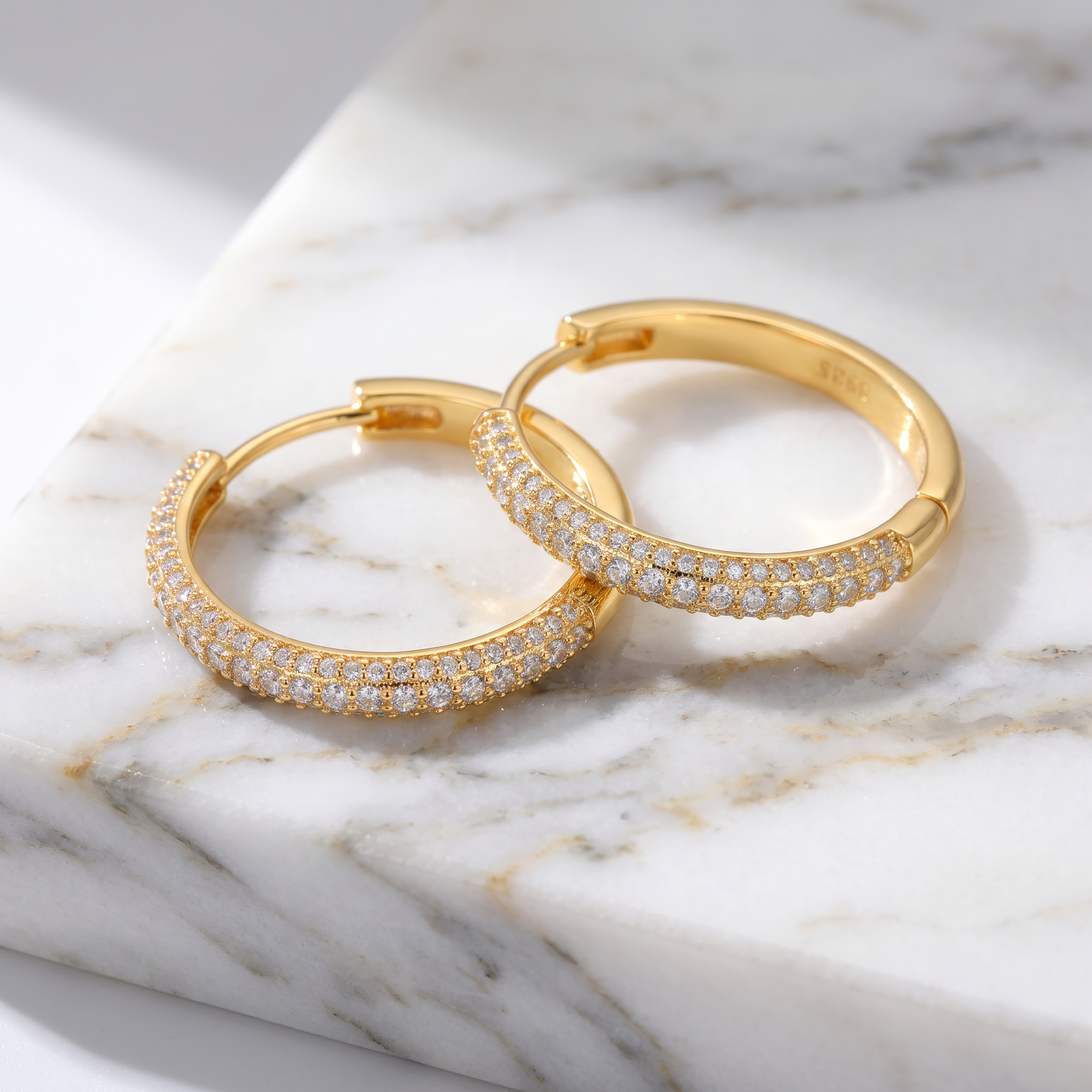 A pair of gold hoop earrings paved with small clear stones, resting on a white and gray marble surface.