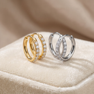 Two triple-hoop huggie earrings with pavé-set clear stones, shown in yellow gold and silver-tone metal, resting on a beige velvet jewelry box.