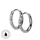A pair of silver hoop earrings with clear pavé-set stones on one side, shown on a white background with a circular 'Skin Safe Waterproof' badge in the lower-left corner.