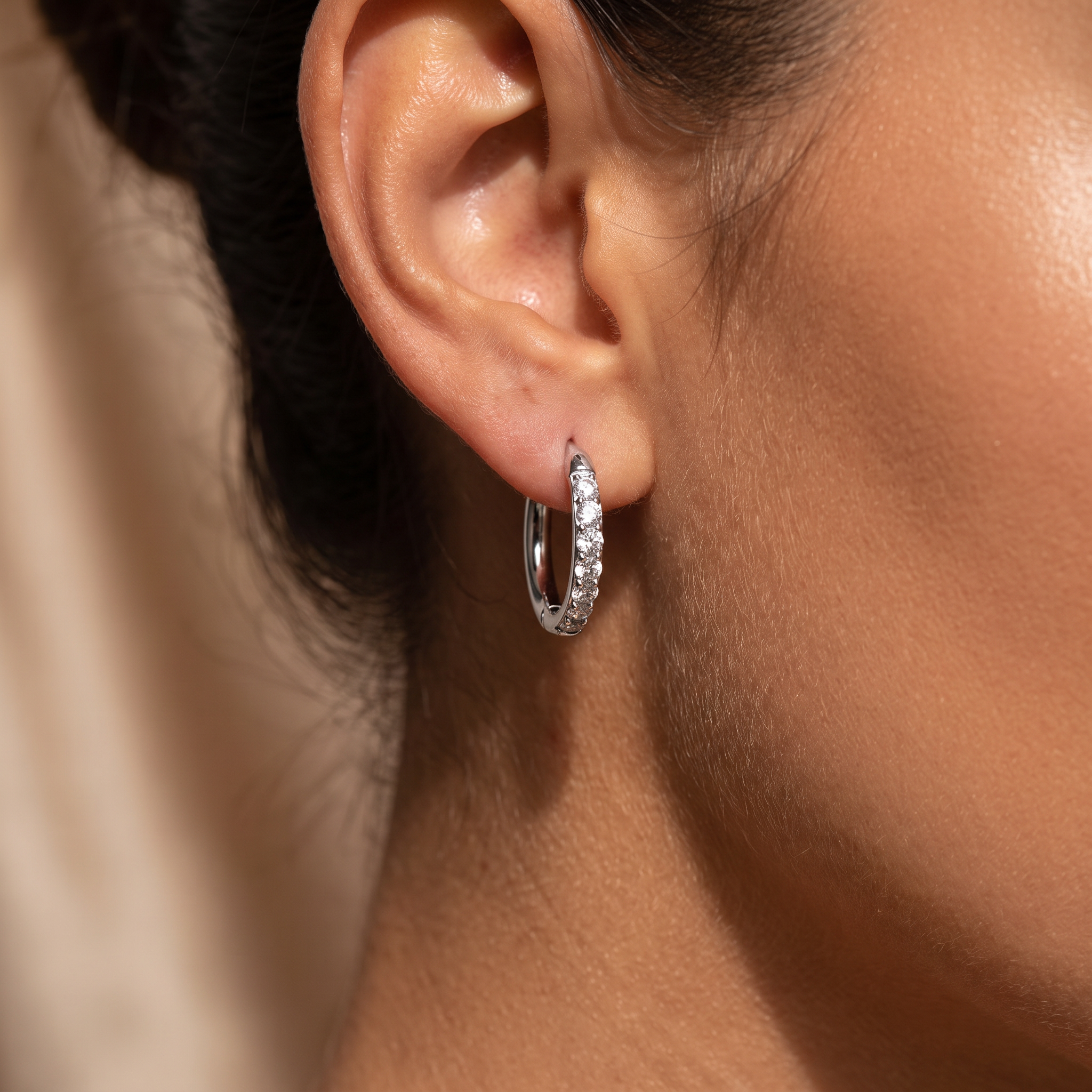 A close-up of a model's ear wearing a small silver hoop earring set with clear stones.