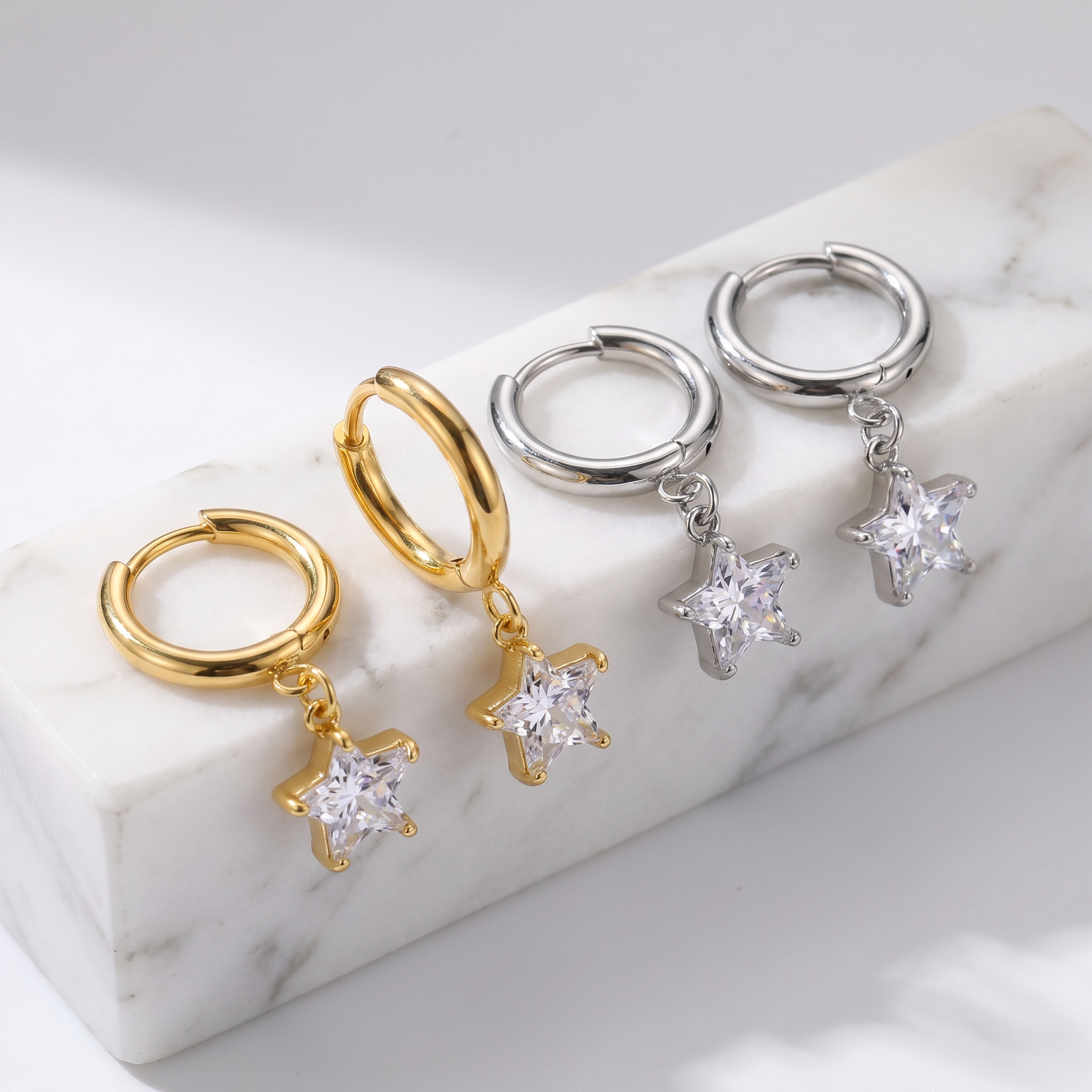 Two pairs of small hoop earrings with dangling star-shaped clear stones, shown on a white marble block. One pair is gold-toned and the other is silver-toned.