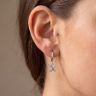 A close-up of a person's ear wearing a silver hoop earring with a dangling clear crystal flower charm, alongside a small silver stud piercing.