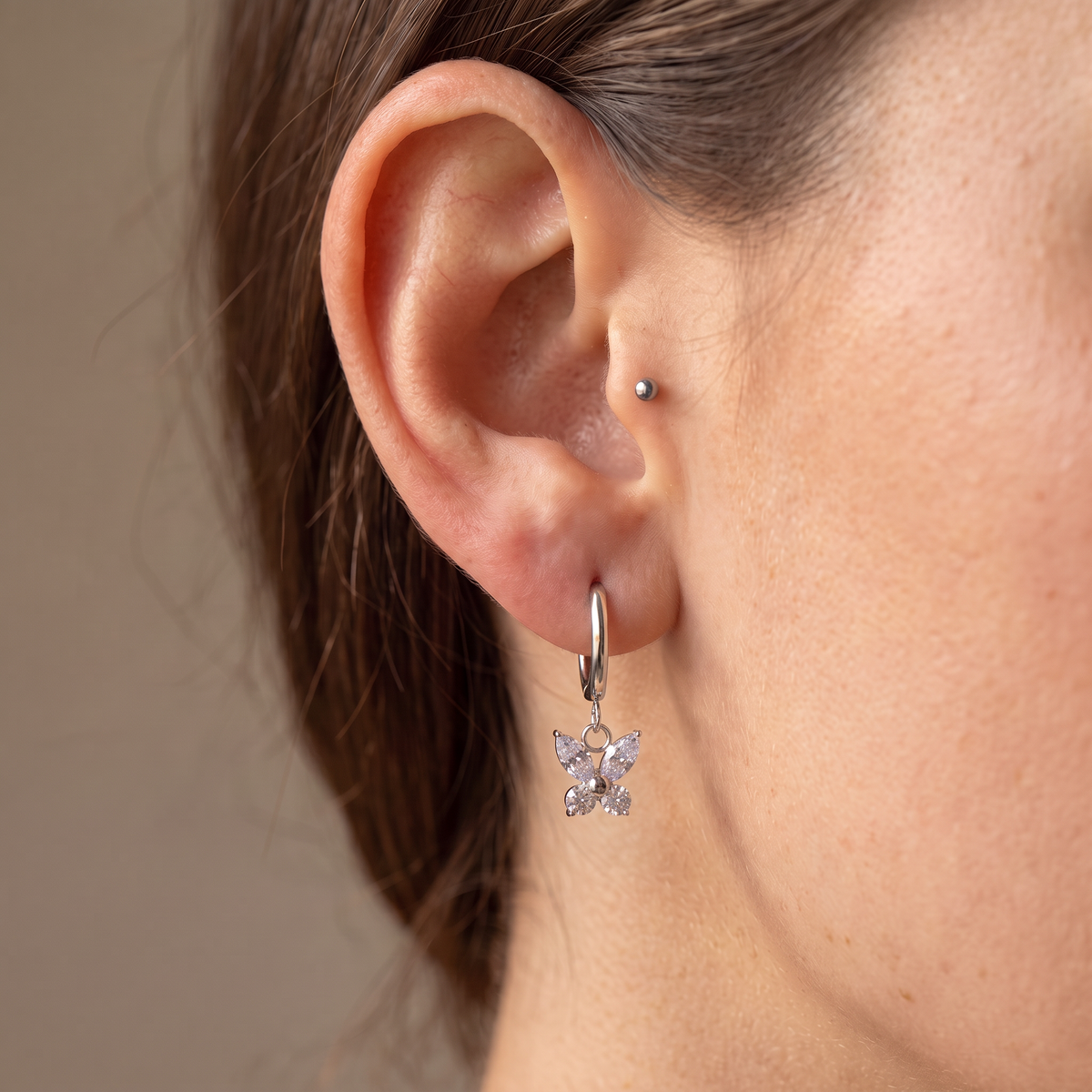 A close-up of a person's ear wearing a silver hoop earring with a dangling clear crystal flower charm, alongside a small silver stud piercing.
