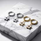 Four small hoop earrings with dangling flower charms are arranged on a white marble block. Two pairs are silver-toned and two are gold-toned, each with clear stones in the flower center.