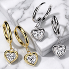 Three pairs of hoop-and-heart drop earrings arranged on a white marble surface: two gold-tone and one silver-tone, each with a heart-shaped clear stone dangling below the hoop.