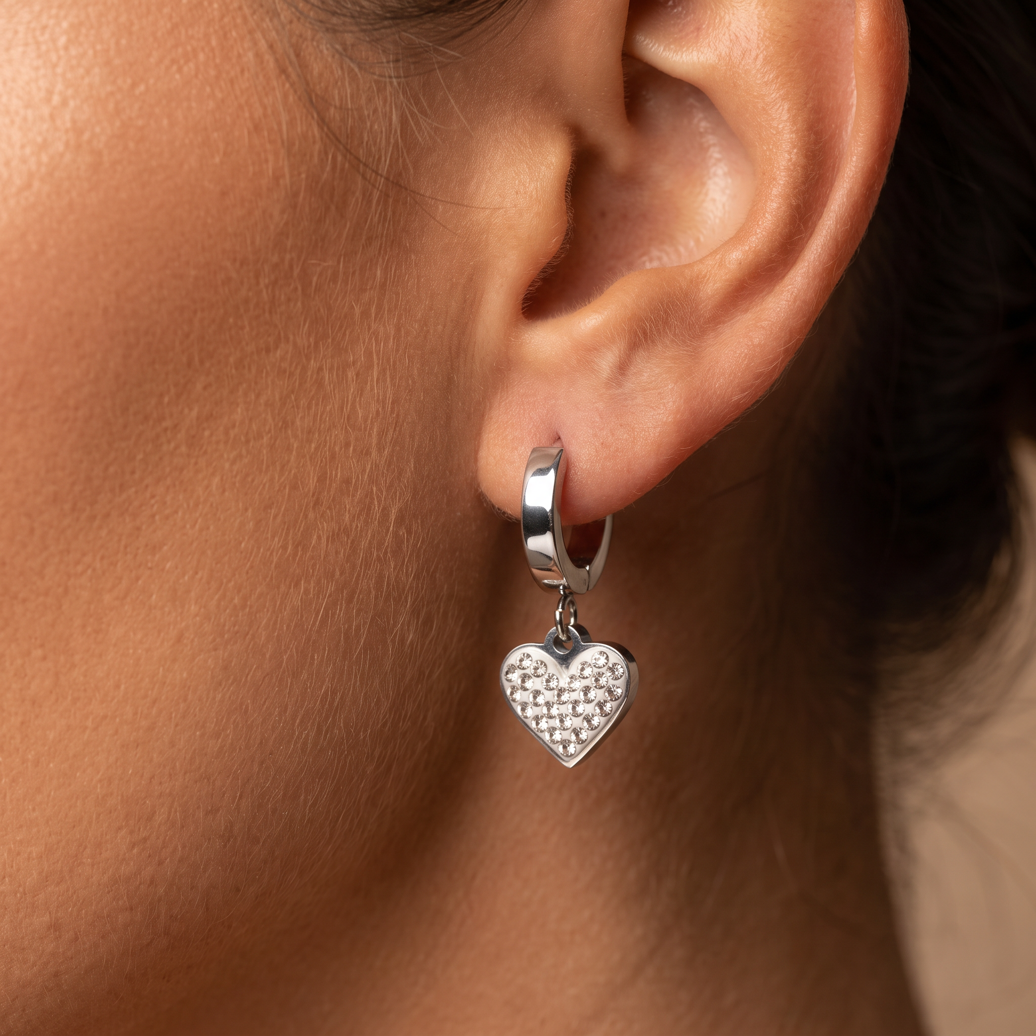 A close-up of a person's ear wearing a silver huggie hoop earring with a dangling textured heart charm.