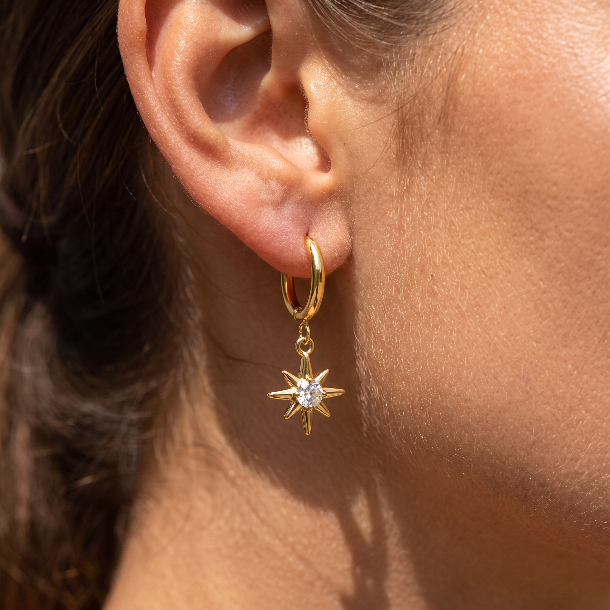 A close-up of a person's ear wearing a gold huggie hoop earring with a dangling gold compass star charm set with a clear gemstone.