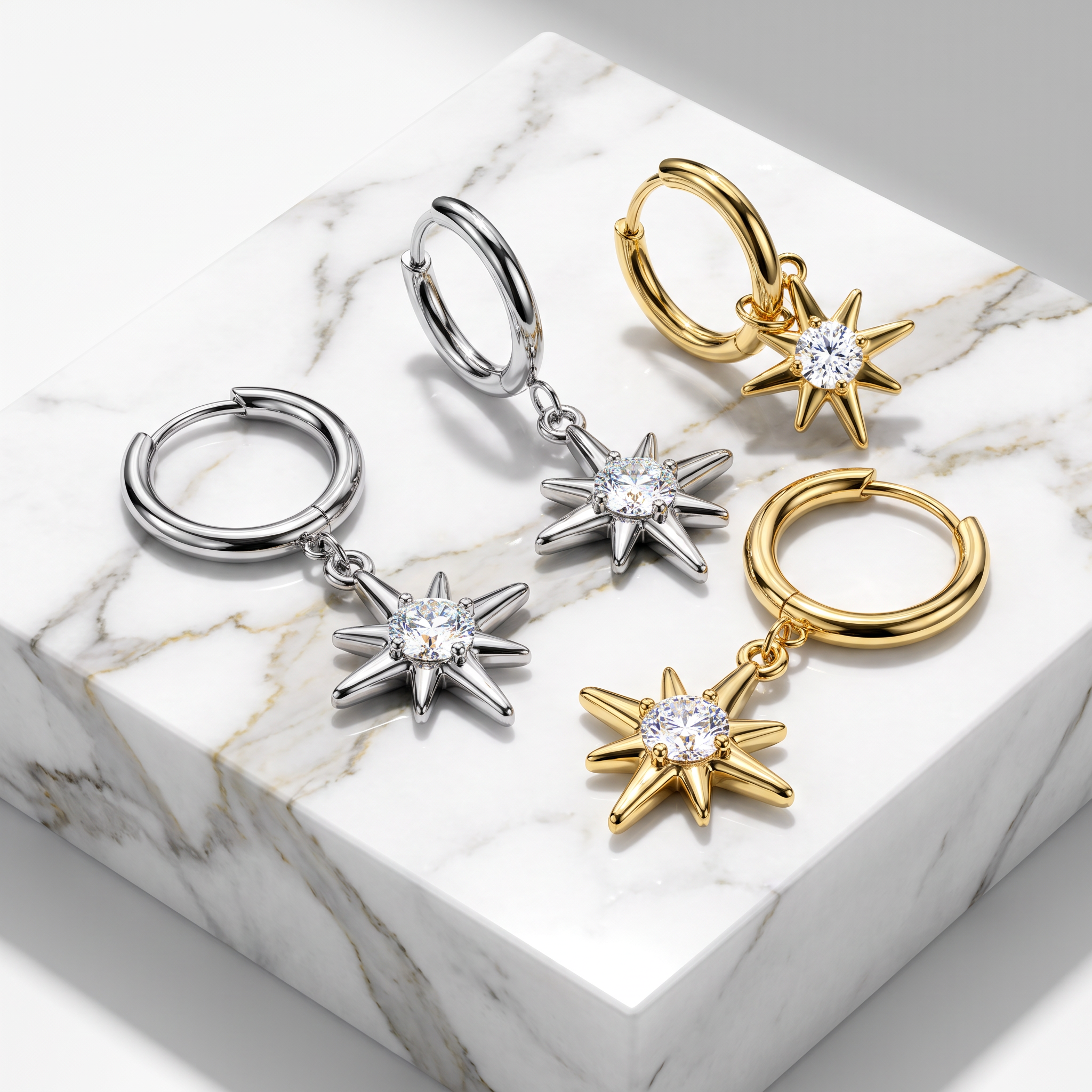 Four small hoop earrings with dangling starburst charms are arranged on a white marble block. Two hoops are silver-toned and two are gold-toned, each with a clear faceted stone in the center of the starburst charm.