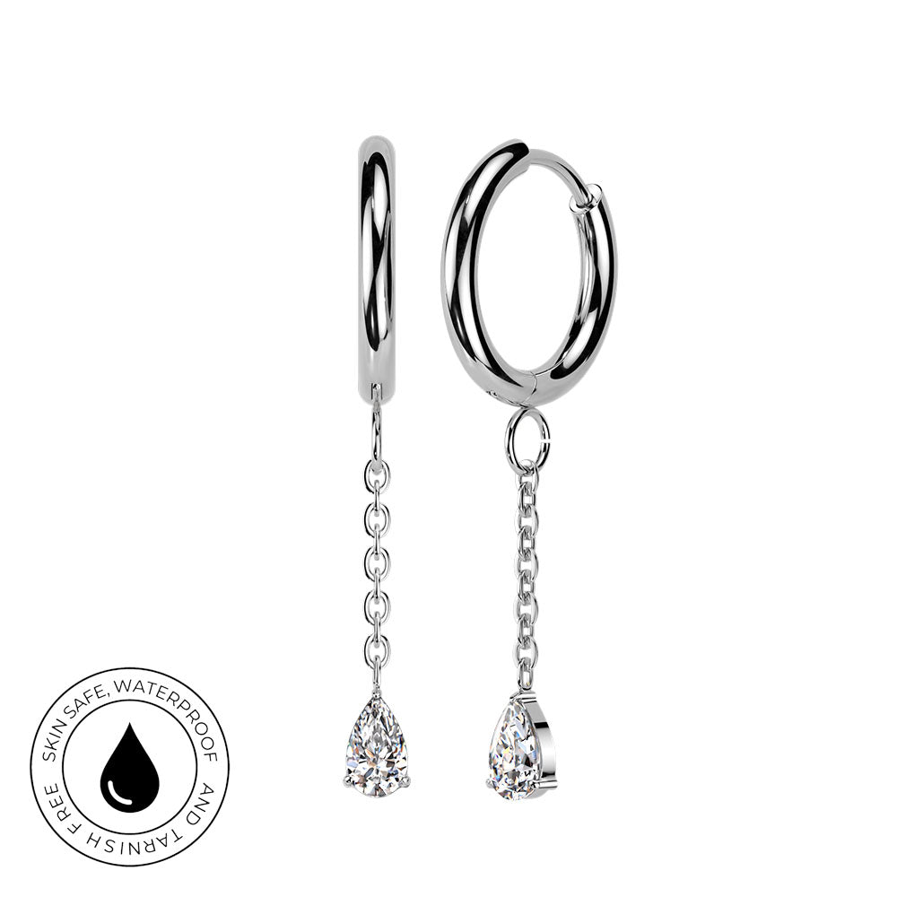 A pair of silver huggie hoop earrings with dangling chains and teardrop-shaped clear gemstones, shown on a white background with a circular 'Skin Safe Waterproof' badge in the lower left corner.