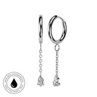 A pair of silver huggie hoop earrings with dangling chains and teardrop-shaped clear gemstones, shown on a white background with a circular 'Skin Safe Waterproof' badge in the lower left corner.