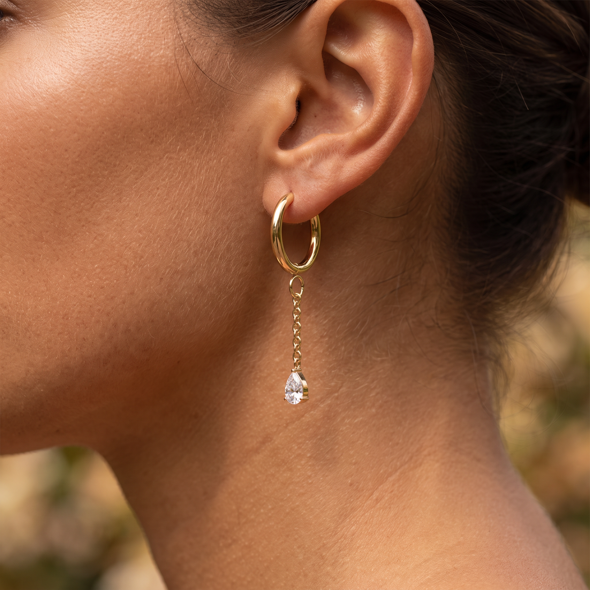 A close-up of a model's ear wearing a gold hoop earring with a delicate chain and a teardrop-shaped clear gemstone dangle.