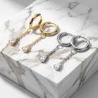 Two pairs of dangling hoop earrings displayed on a white marble block. One pair is yellow gold with a small teardrop-shaped clear gemstone at the bottom of each hoop and chain.