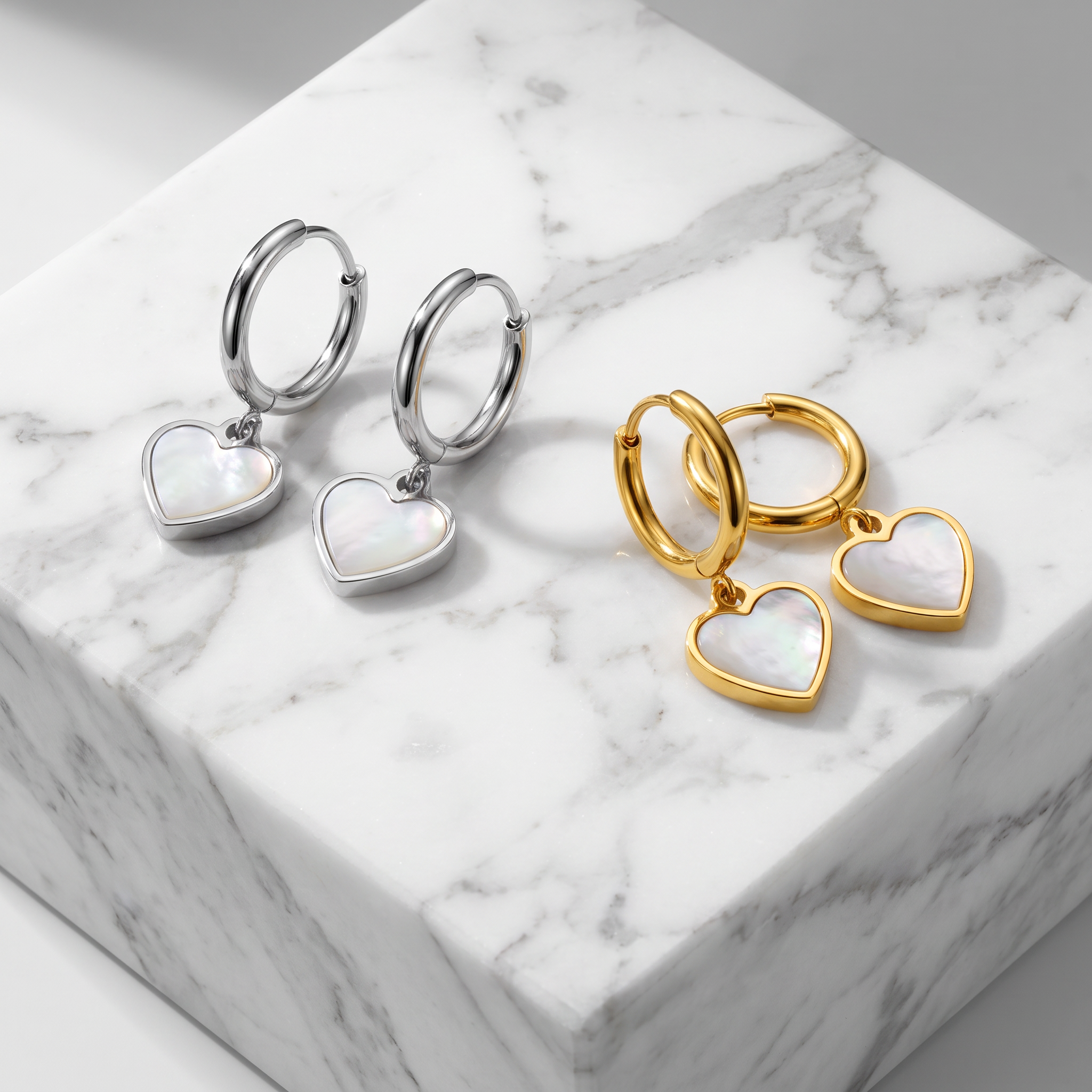 Two pairs of small hoop earrings with heart-shaped pendants are arranged on a white marble block. One pair is silver-toned with iridescent white heart charms, and the other is gold-toned with matching heart charms.