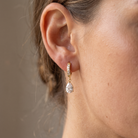 A close-up of a person's ear wearing a small gold huggie hoop earring adorned with clear stones and a dangling pear-cut clear gemstone.