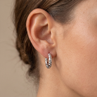 A close-up of a person's ear wearing a small silver hoop earring with a textured, hammered finish against a neutral beige background.