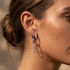 A close-up of a woman's ear wearing a silver hoop earring with a dangling textured chain detail, set against a softly blurred warm-toned background.
