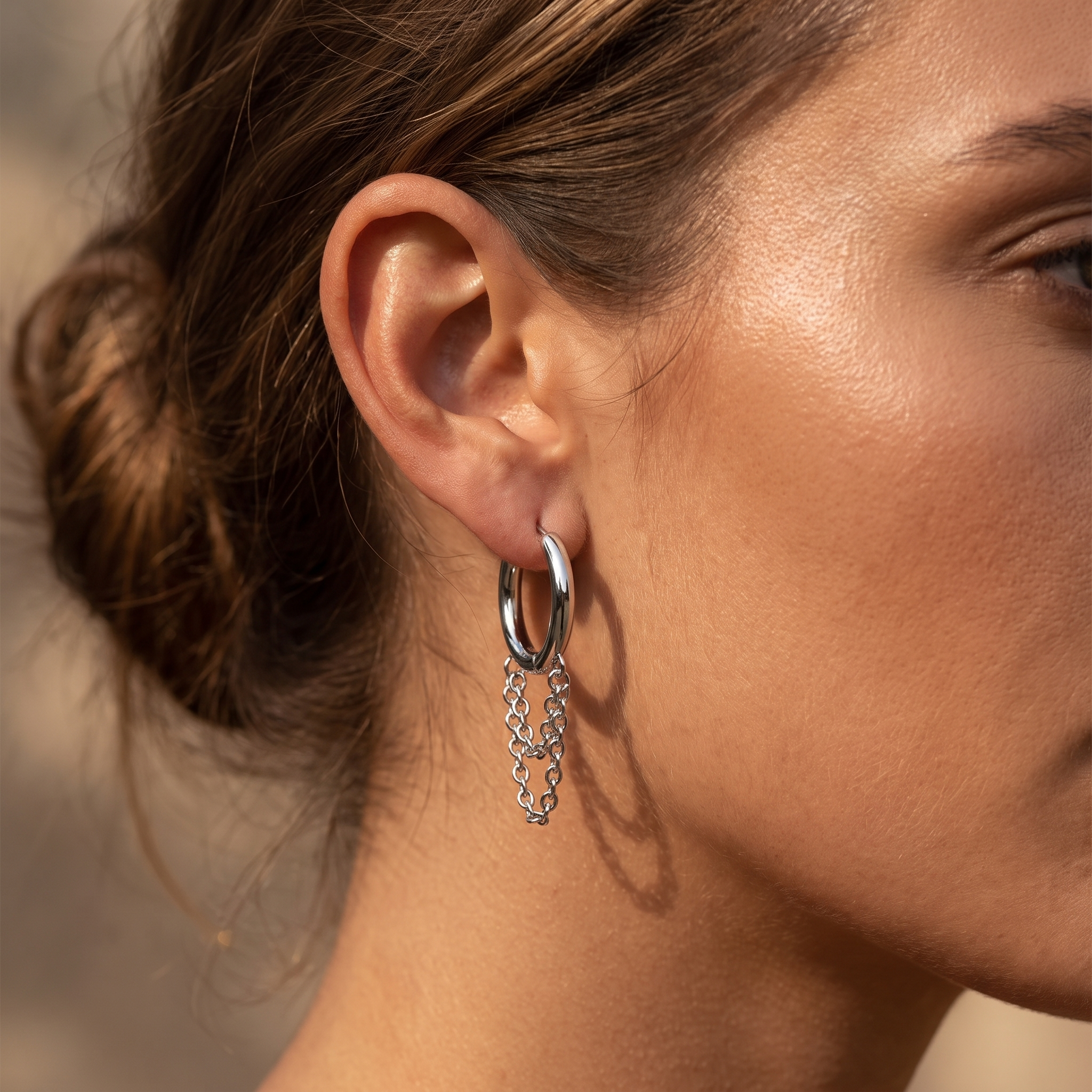 A close-up of a woman's ear wearing a silver hoop earring with a dangling textured chain detail, set against a softly blurred warm-toned background.