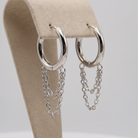 Silver hoop earrings with dangling chain links, displayed on a beige jewelry stand against a light gray background.