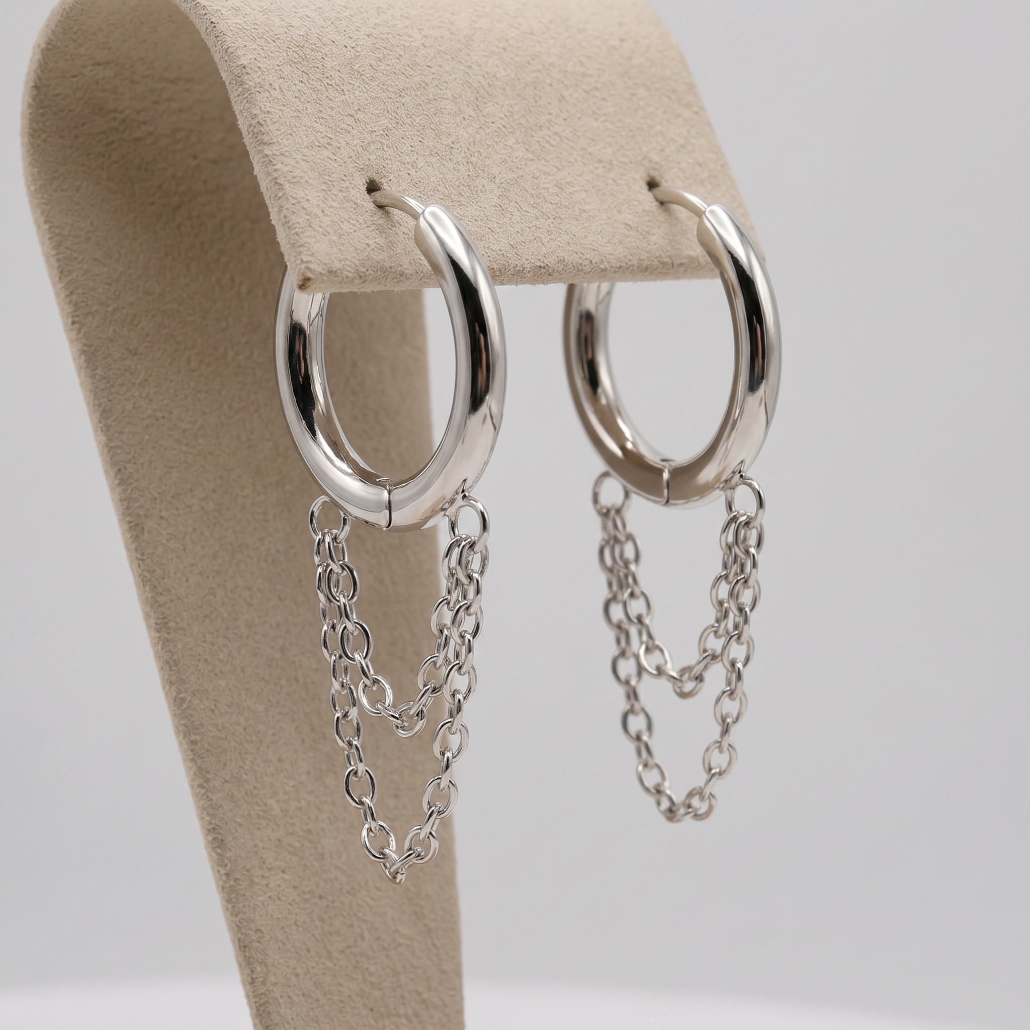 Silver hoop earrings with dangling chain links, displayed on a beige jewelry stand against a light gray background.