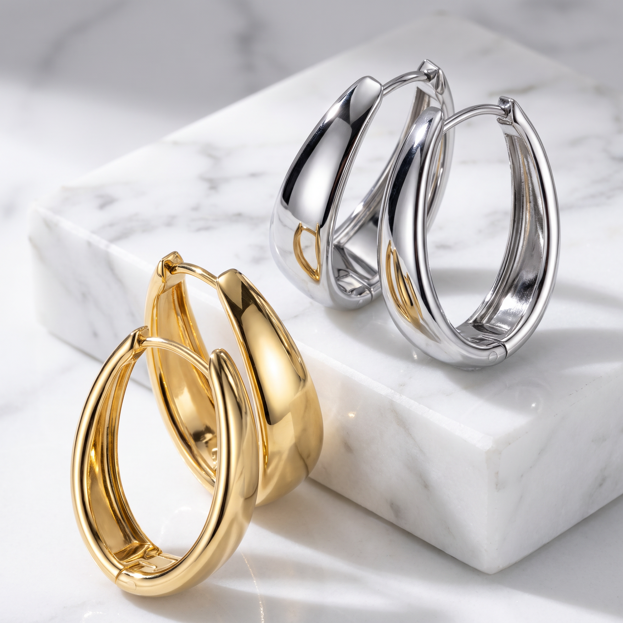 Two pairs of polished hoop earrings, one in yellow gold and one in silver, displayed on a white marble block over a light marble surface.