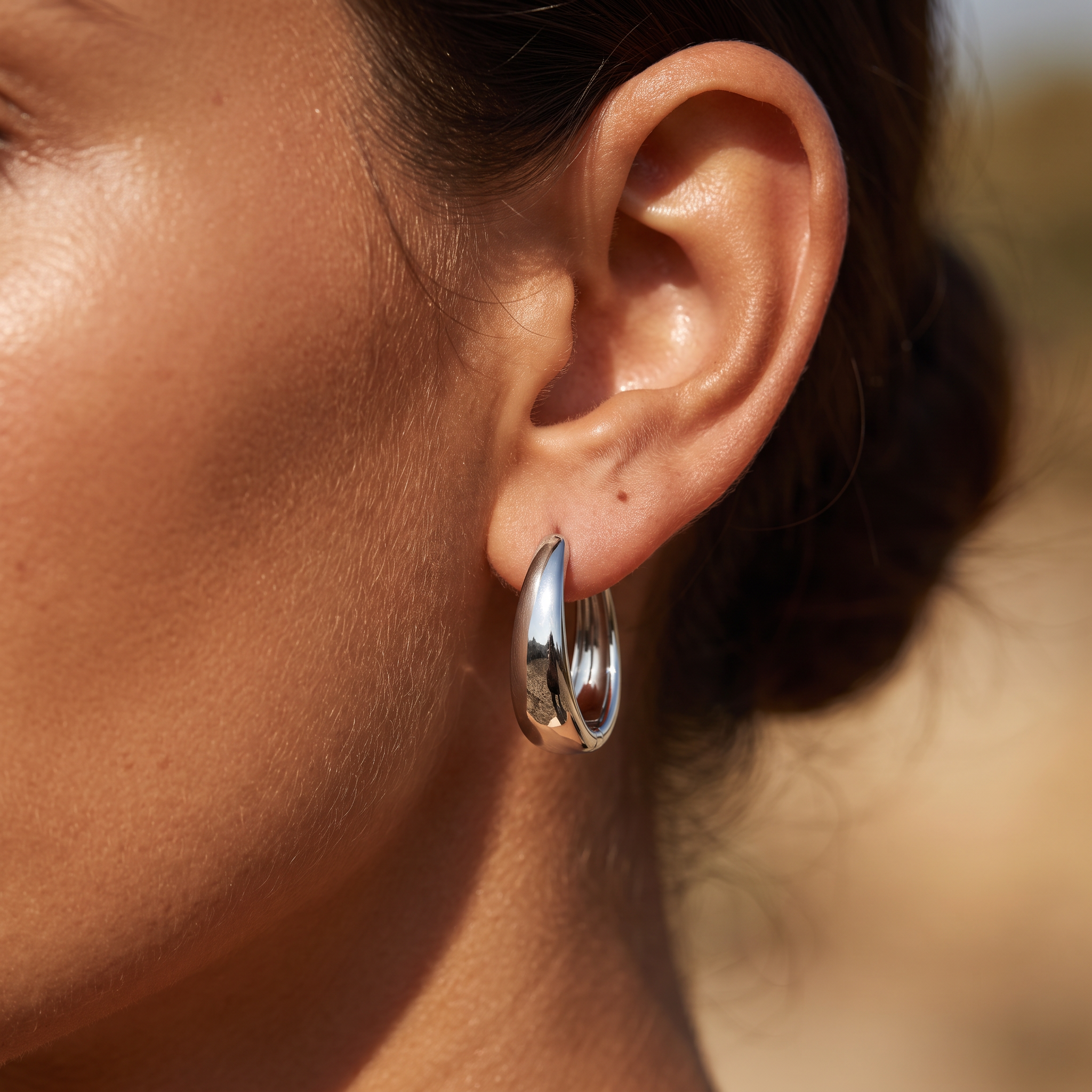 A close-up of a person's ear wearing a polished silver hoop earring in warm natural light.