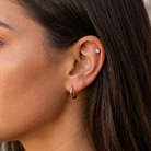 Close-up of a person's ear wearing a small gold hoop earring and a tiny round stud earring, with dark brown hair and warm skin tones.