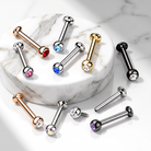 Multiple barbell-style body jewelry piercings with gemstone accents are arranged on and around a round white marble pedestal against a white marble surface. The pieces are shown in rose gold, silver, gold, black, red, blue, pink, purple, and iridescent finishes.