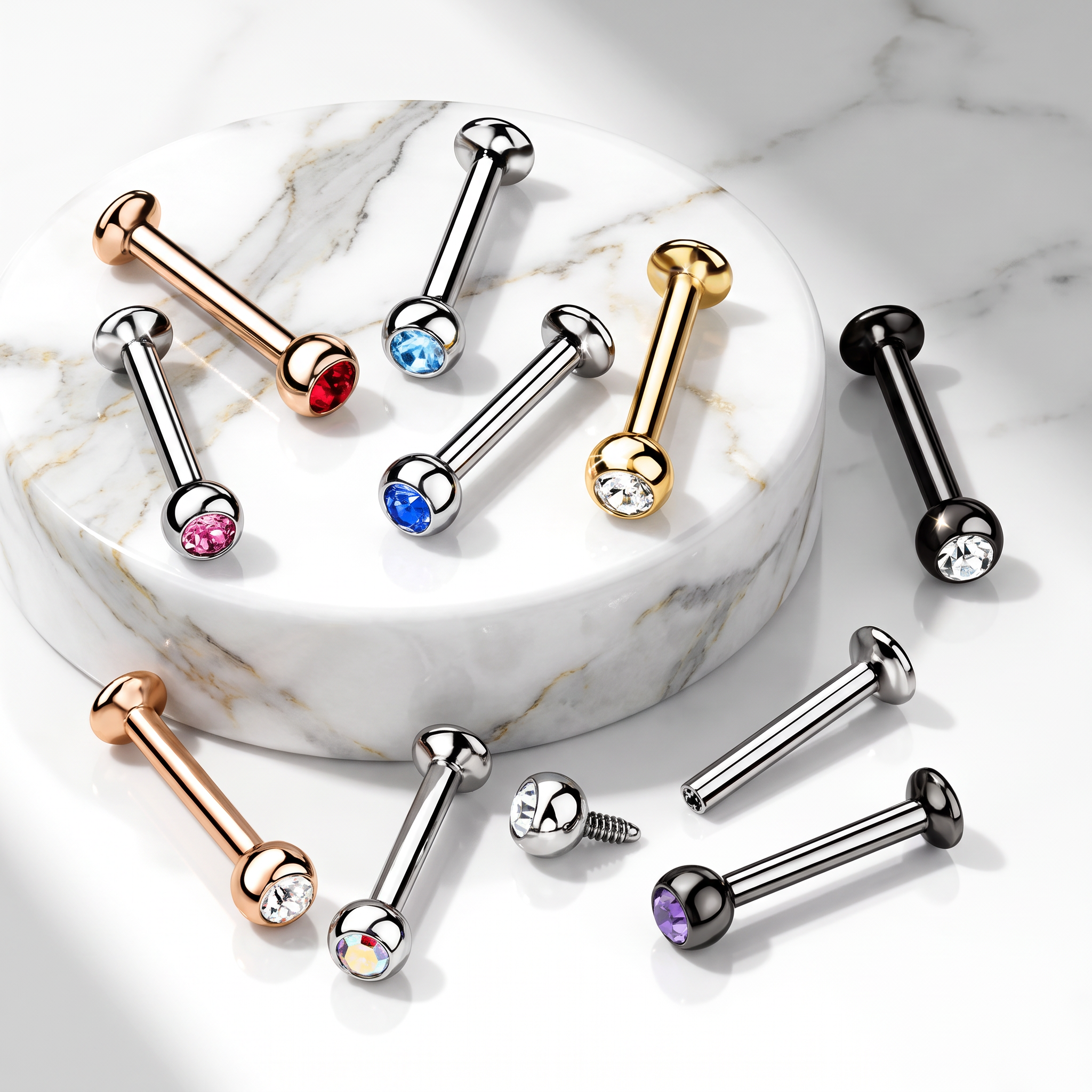 Multiple barbell-style body jewelry piercings with gemstone accents are arranged on and around a round white marble pedestal against a white marble surface. The pieces are shown in rose gold, silver, gold, black, red, blue, pink, purple, and iridescent finishes.