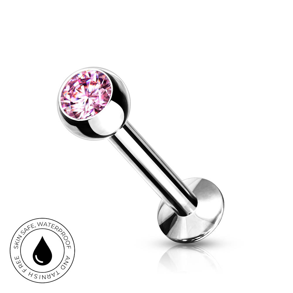 A silver-toned labret piercing barbell with a faceted pink gemstone ball end, shown on a clean white background with a small circular skin-safe waterproof badge in the lower left corner.