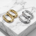 Two pairs of chunky capsule-shaped hoop earrings, one in polished gold and one in polished silver, displayed on a white marble block.