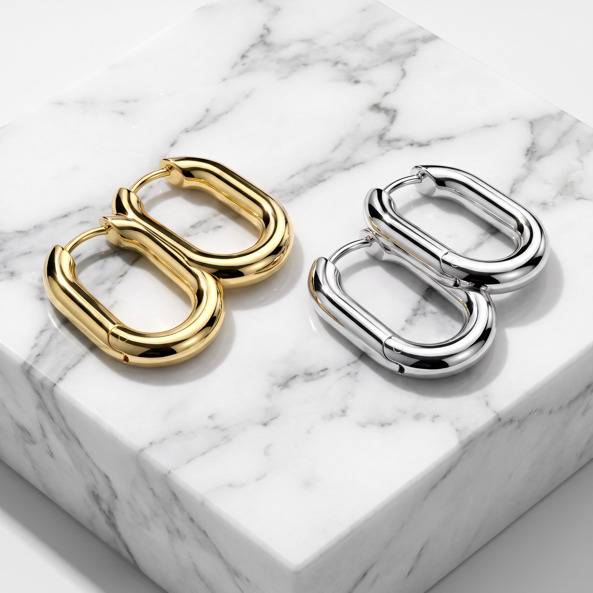 Two pairs of chunky capsule-shaped hoop earrings, one in polished gold and one in polished silver, displayed on a white marble block.