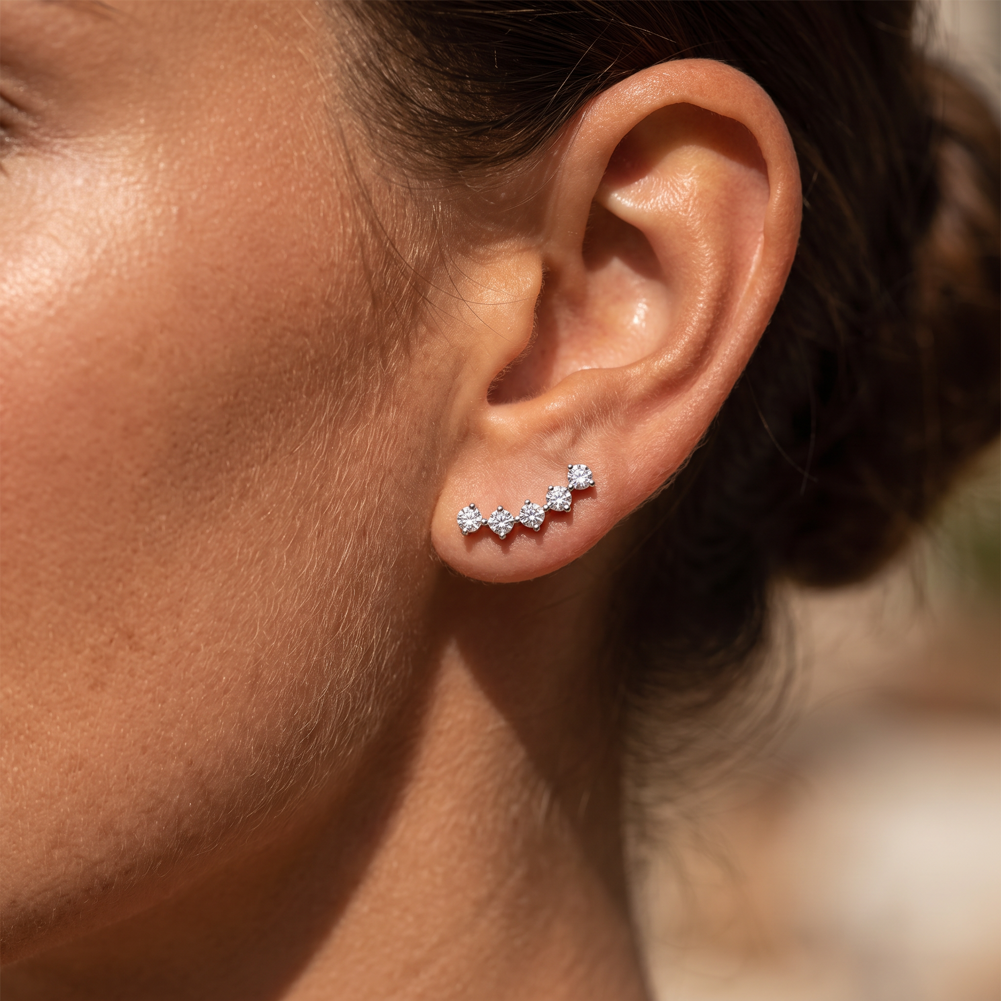 A close-up of a woman's ear wearing a small cluster of silver stud earrings with clear stones, set against a softly blurred warm-toned background.