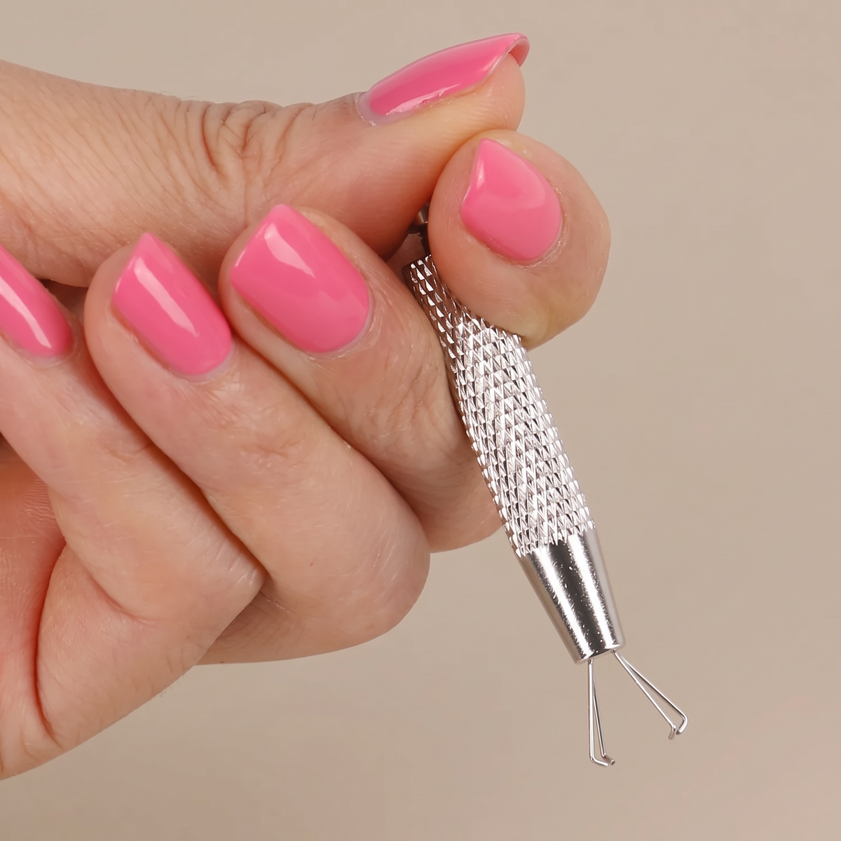 A close-up of a hand with pink manicured nails holding a silver metal nail art tool with a knurled handle and two small curved tips.